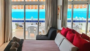 A hotel room with a large sliding glass door leading to a balcony with a view of the sea. There is a bed with red and white pillows, a chair, and a small table with a fan. - Azahara III Studio, Playa Las Vistas with wonderful seaview (Playa de la Américas)
