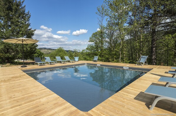A rectangular swimming pool with a wooden deck, surrounded by lounge chairs and a large umbrella.