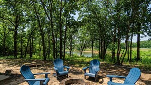 Un camping con cuatro sillas Adirondack dispuestas alrededor de un brasero de piedra, rodeado de árboles altos y un lago al fondo.