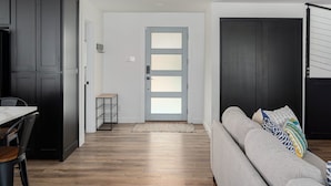 Una cocina moderna con muebles oscuros, un comedor con mesa blanca y un salón con sofá gris.