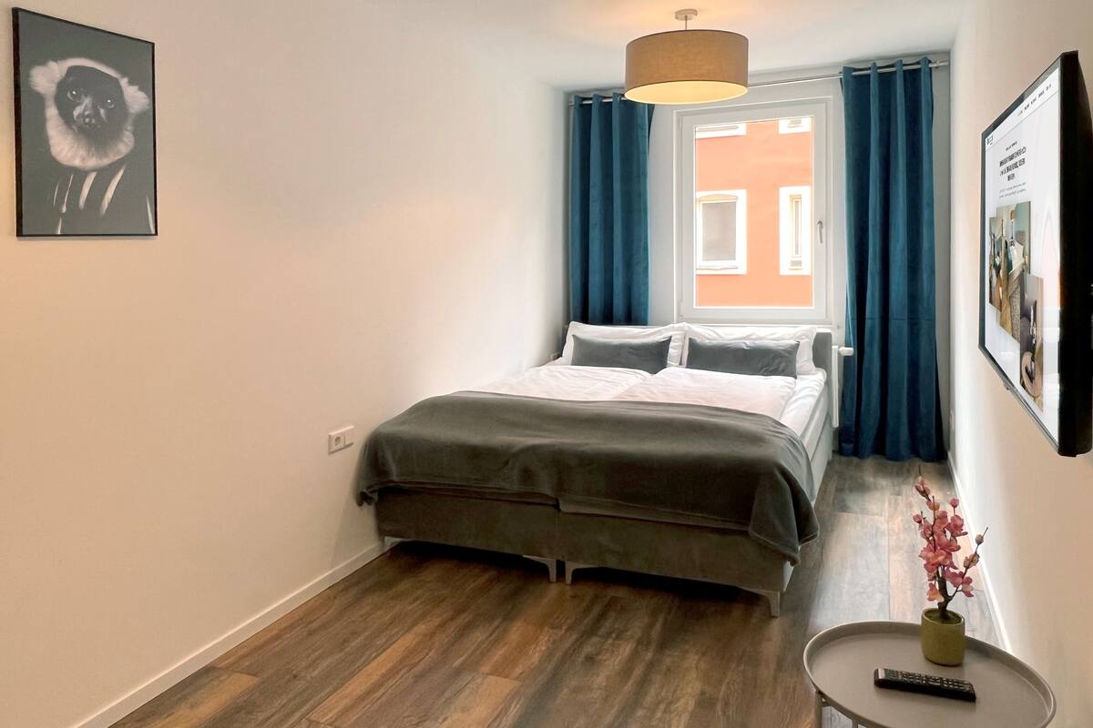 Une chambre à coucher avec un lit, une table de chevet avec une télécommande, un tableau encadré au mur et une fenêtre avec des rideaux.