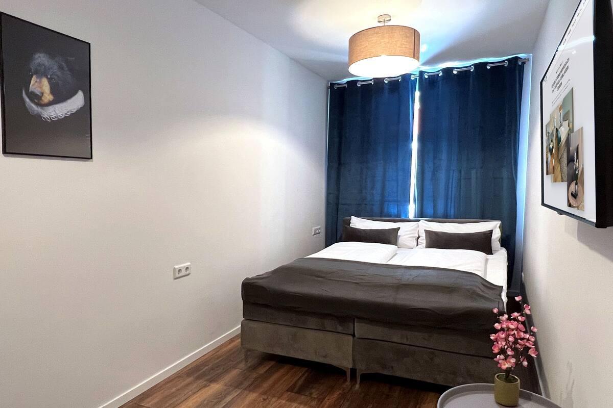 Une chambre à coucher avec un lit, une petite table sur laquelle se trouve une télécommande, une plante en pot et un tableau encadré accroché au mur.