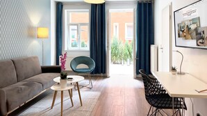 A modern living room with a sofa, a round table, a blue chair, and a desk with a chair. There is a window with curtains, a lamp, and a framed picture on the wall. - Nurban Apartments City 2 (Nuremberg)