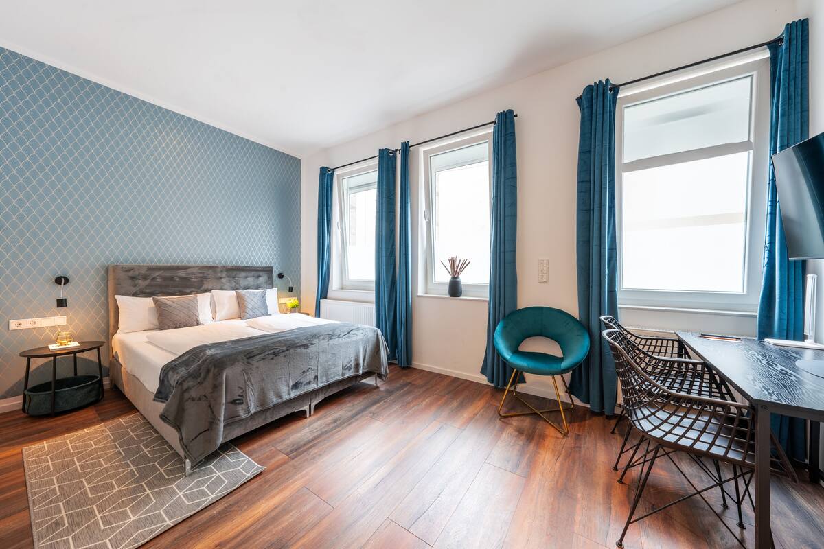 Une chambre à coucher moderne avec un lit, un bureau et une chaise. On y trouve des fenêtres avec des rideaux, une table de chevet avec une lampe et un tapis sur le sol.