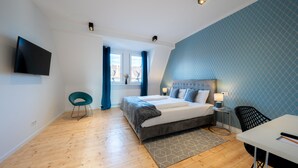 A modern bedroom with a large bed, a TV mounted on the wall, a desk with a chair, and a window with curtains. - Nurban Apartments City 2 (Nuremberg)