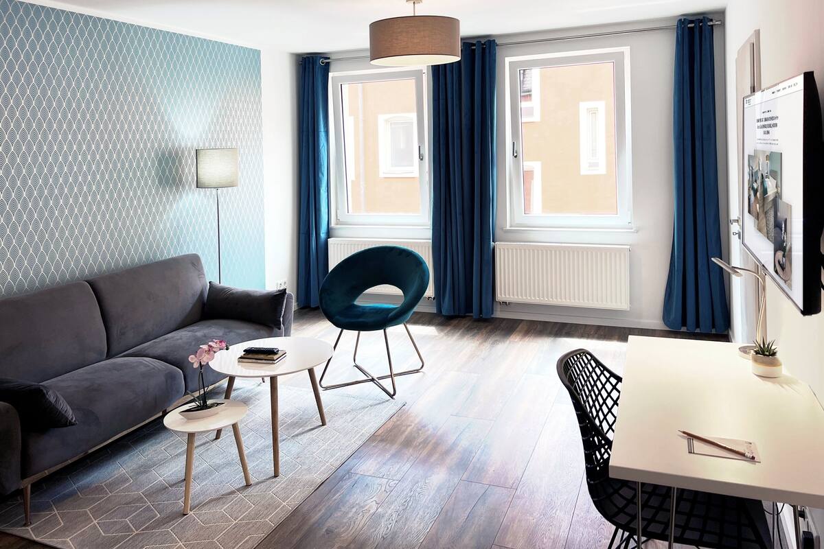 Un salon moderne comprenant un canapé, une table ronde, une chaise et une table à manger. Il y a deux fenêtres avec des rideaux, un radiateur et une lampe fixée au mur.