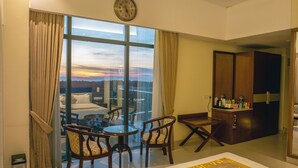 Ein Hotelzimmer mit großem Fenster, einem Bett, einem Esstisch mit Stühlen, einer kleinen Küchenzeile und Blick auf die Stadtlandschaft.