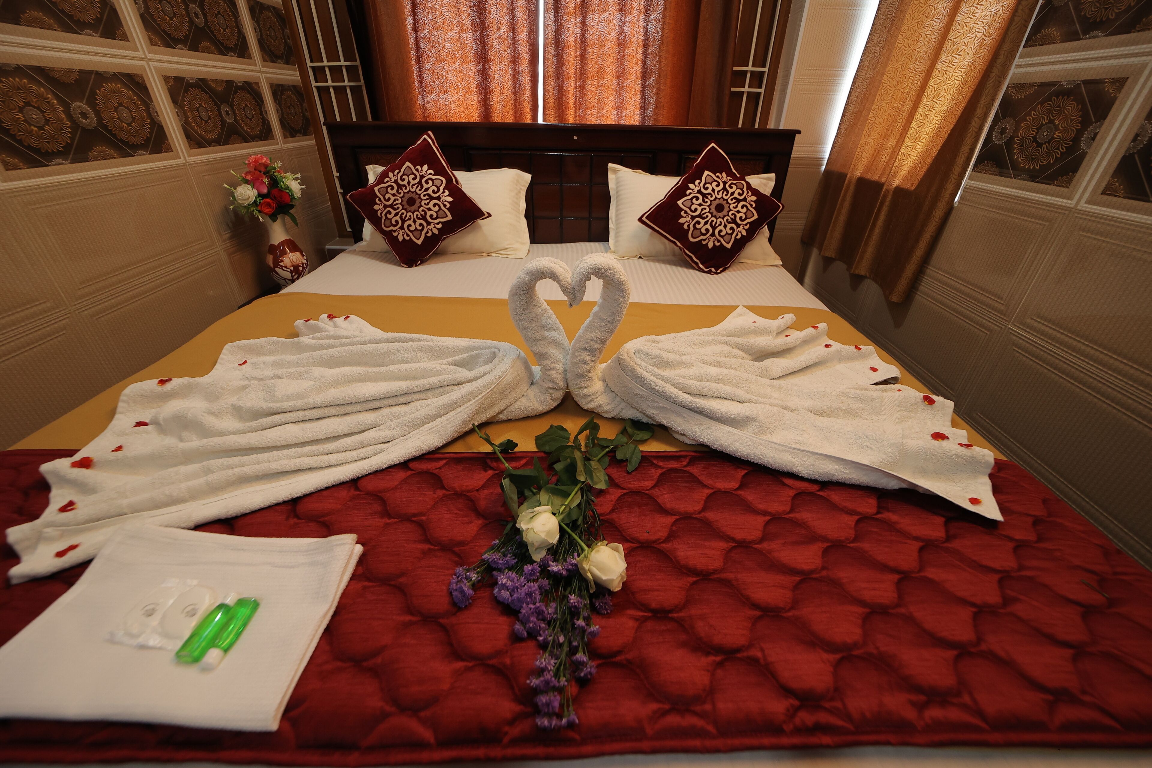A neatly made bed with towels arranged in a heart shape, a bouquet of flowers, and a bottle of lotion on the bed.