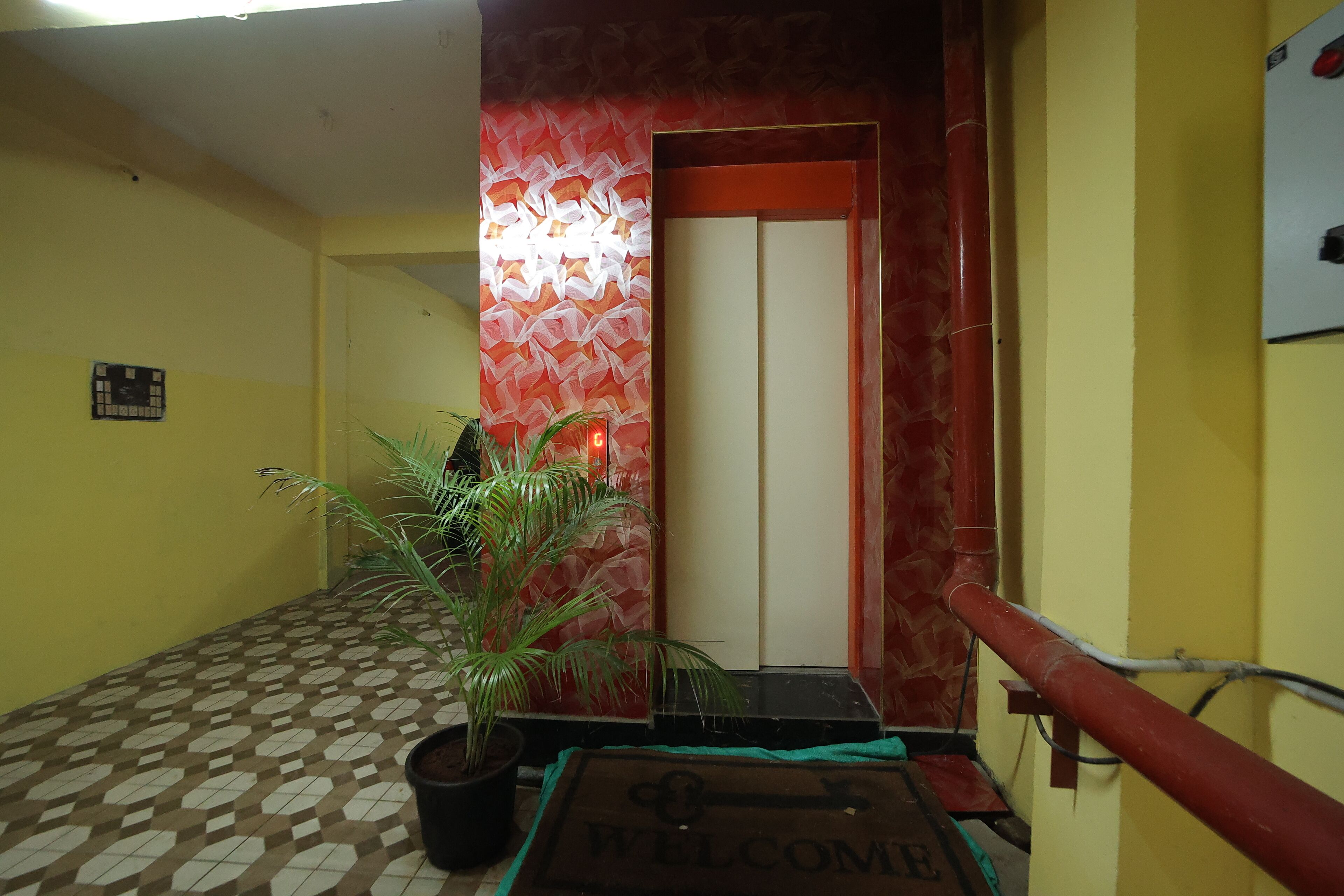 A hallway with a patterned floor, a welcome mat, a potted plant, and a door with a red frame.