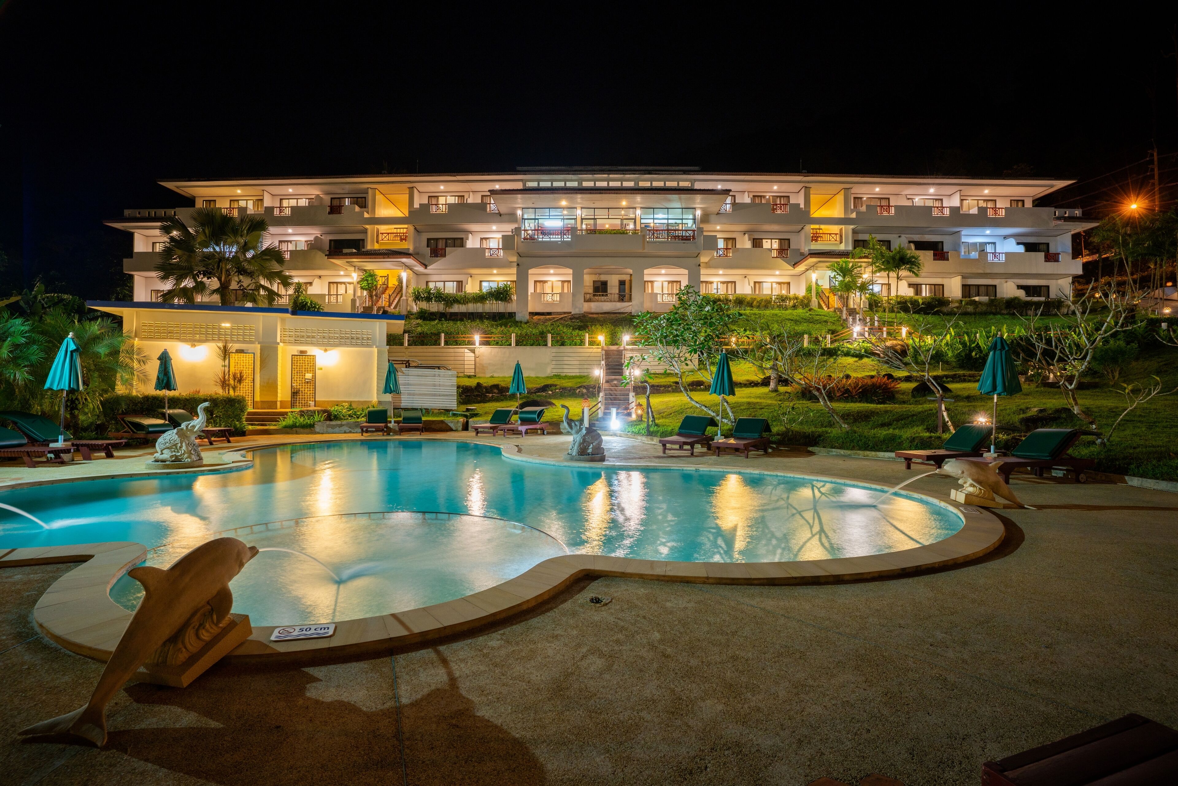 Un hôtel moderne doté d’une piscine, de chaises longues et d’un espace extérieur bien éclairé la nuit.
