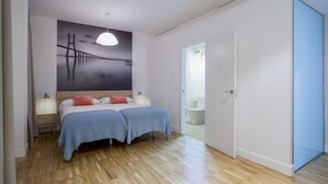 A bedroom with two beds, a nightstand, a wall-mounted lamp, and a door leading to a bathroom.