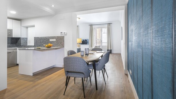 A modern kitchen with a granite countertop, a dining area with a wooden table and patterned chairs, and a blue textured wall.