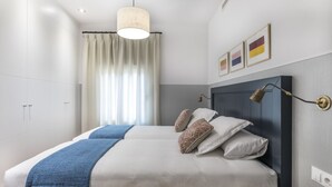 A bedroom with two beds, a large window with curtains, a ceiling light, and a wall-mounted lamp.