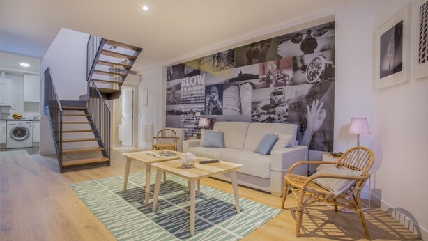 Ein modernes Wohnzimmer mit Treppe, einer großen Wandmalerei, einem Sofa, einem Couchtisch und einem Korbstuhl.