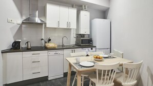 A compact kitchen with white cabinets, a stainless steel range hood, a microwave, and a refrigerator. There is a dining table with chairs and a small breakfast setup. - Apartamento Bajo Interior 2 Dorm, 3 Camas (Madrid)