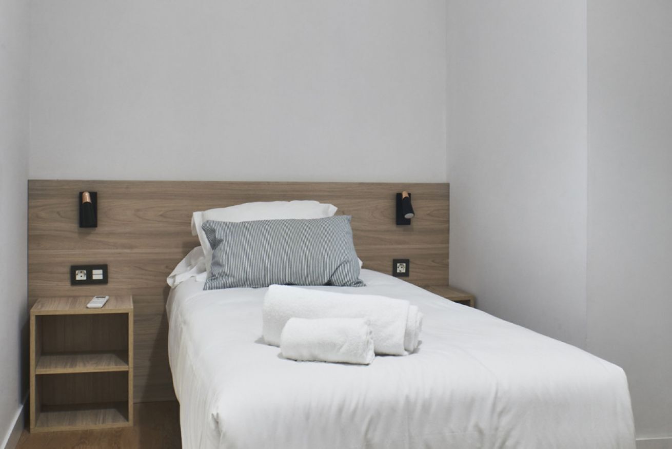 A neatly made bed with white bedding and a gray striped pillow, a wooden nightstand with a phone, and a wall-mounted light fixture.