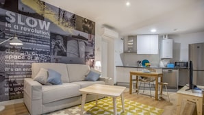 Een moderne woonkamer met een bank, salontafel en een gekleurd tapijt. Achtergrond toont een keuken met witte kasten en apparatuur.