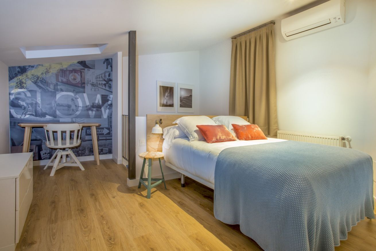 Ein Schlafzimmer mit einem Bett, einem Schreibtisch und einem Stuhl. Ein Fenster mit Vorhängen, eine Klimaanlage und eine Wand mit Stadtbild-Mural.