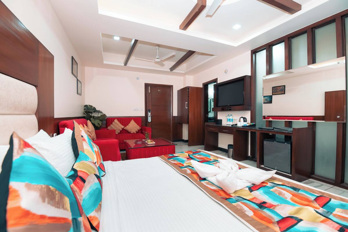 A hotel room with a bed, a red sofa, a desk with a television, and a kitchenette.
