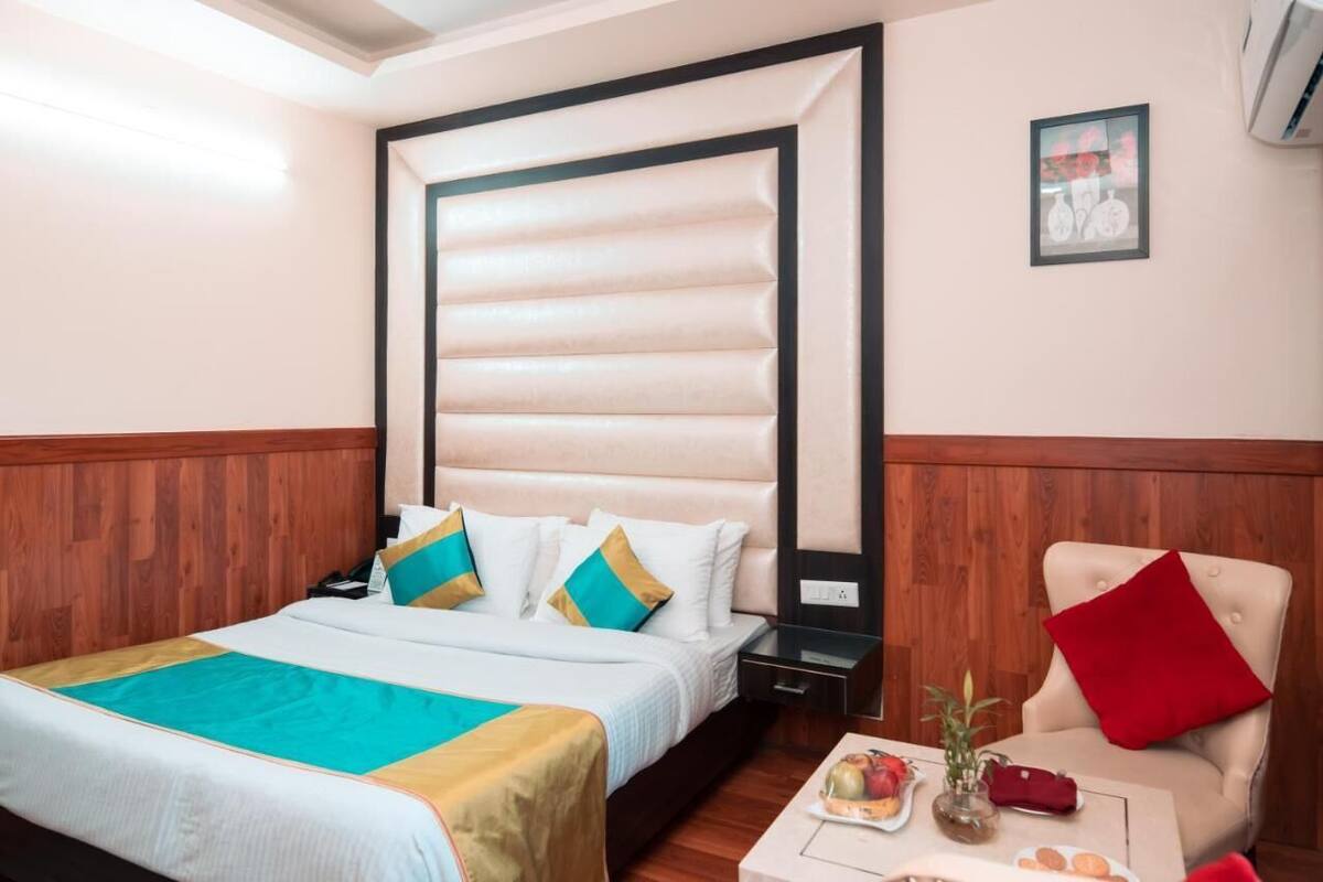 A hotel room with a large bed, a wooden headboard, a small table with a vase and a plate of fruit, and a red pillow on a chair.