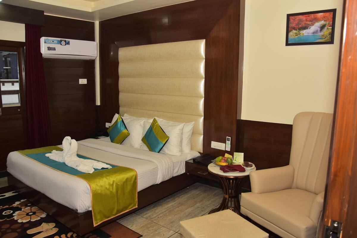 A hotel room with a bed, a chair, a small table with a fruit bowl, and an air conditioning unit.