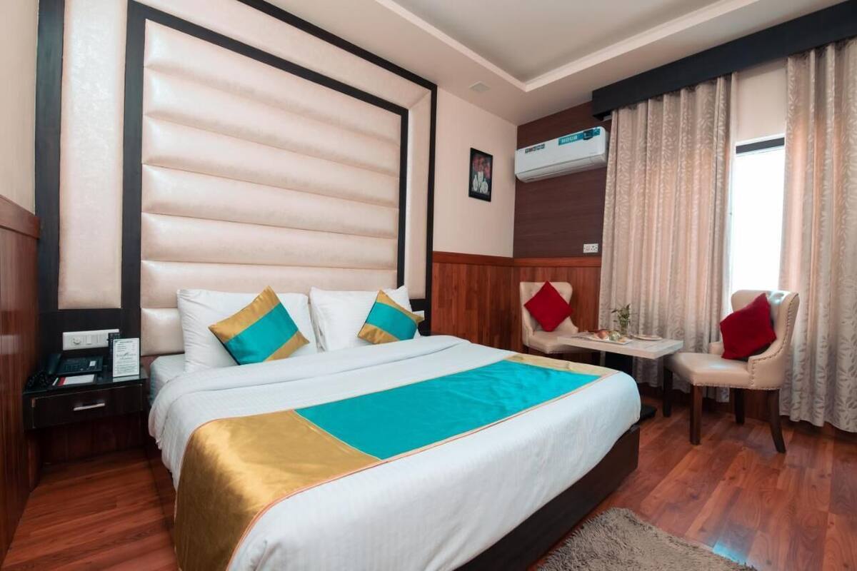 A hotel room with a bed, bedside table, chair, and an air conditioning unit.