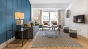 A modern living room with a sofa, coffee table, and ottomans. There is a TV mounted on the wall and a window with curtains. - Apartamento Duplex 2 Dormitorios con Terraza (Madrid)