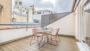 A balcony with a wooden floor, a round table with chairs, and a view of buildings in the background. - Apartamento Duplex 2 Dormitorios con Terraza (Madrid)