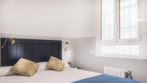Une chambre avec une tête de lit sombre, deux coussins à motifs, une couverture bleue, une lampe de chevet et une fenêtre avec des stores.