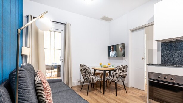 Un salon moderne avec un canapé gris, des coussins à motifs, une table à manger avec des chaises, un téléviseur fixé au mur et un coin cuisine avec une cuisinière.