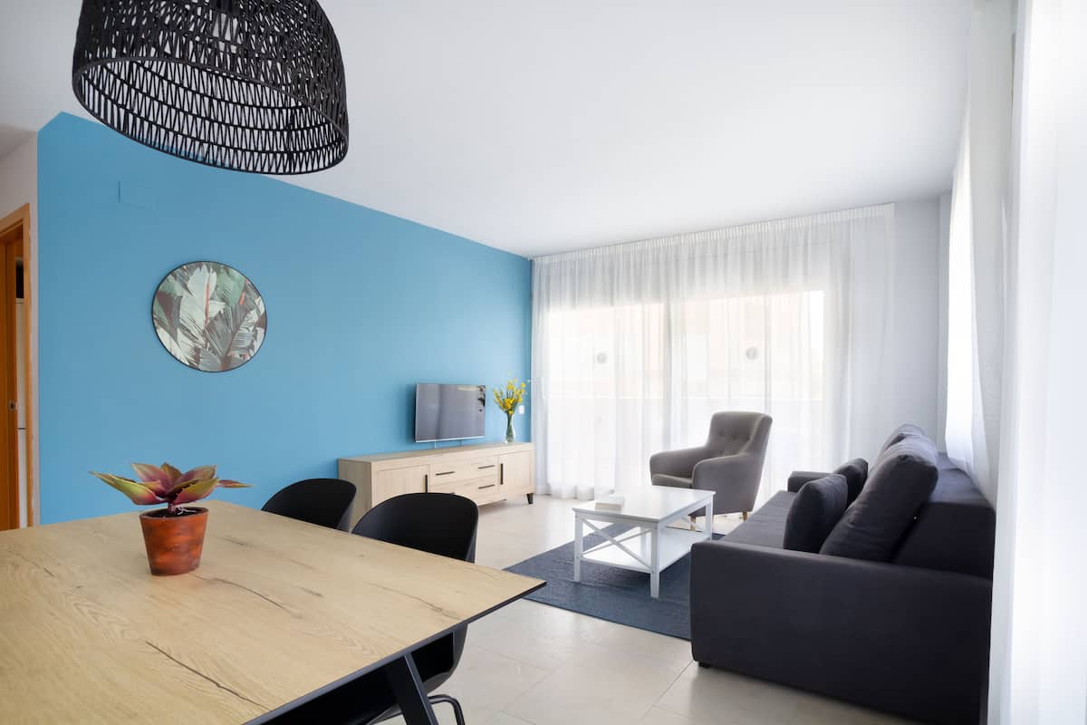 A modern living room with a blue accent wall, a wooden dining table, a grey sofa, a white coffee table, and a TV on a wooden cabinet.