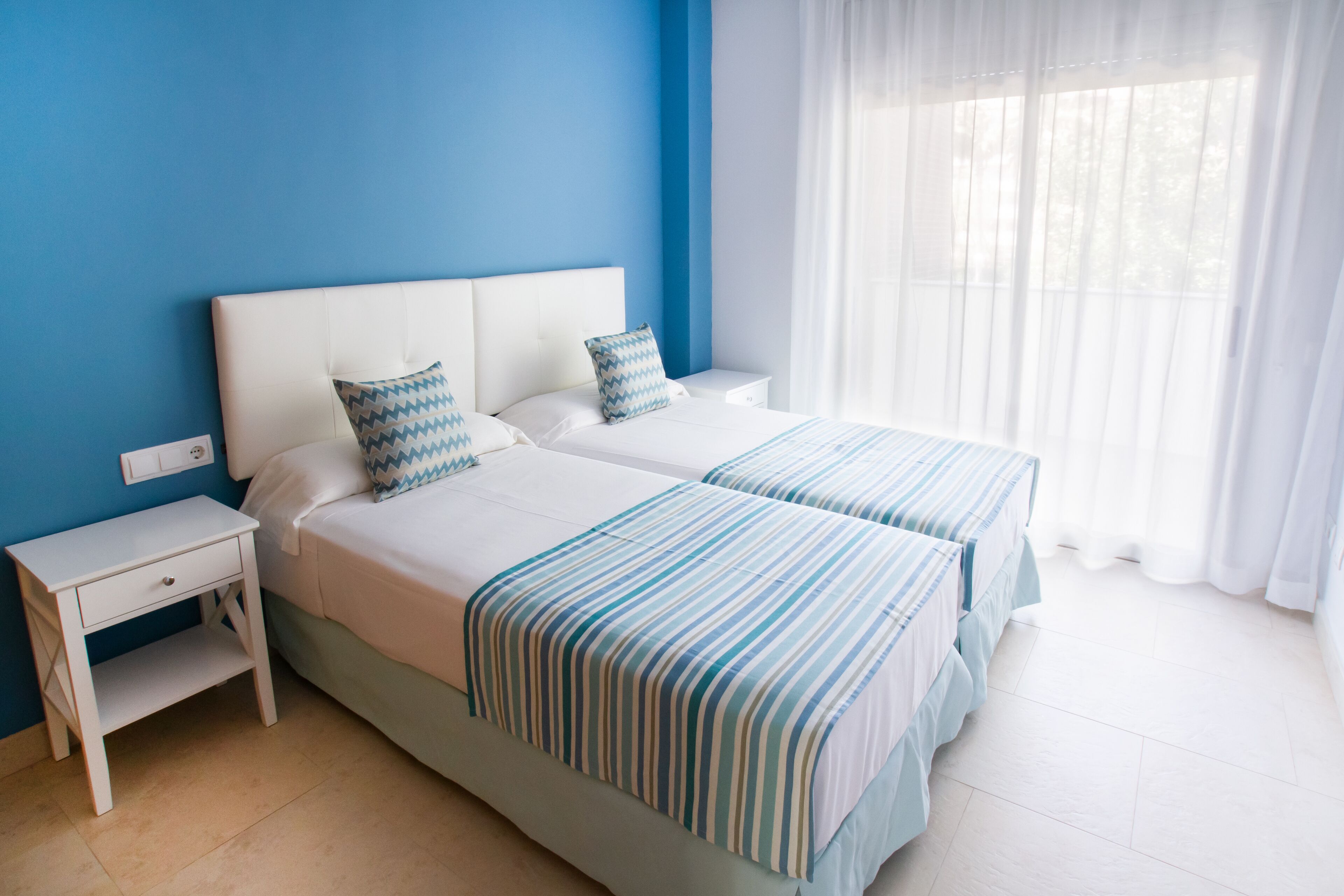 Habitación de hotel con dos camas, una mesita de noche y una ventana con cortinas traslúcidas.