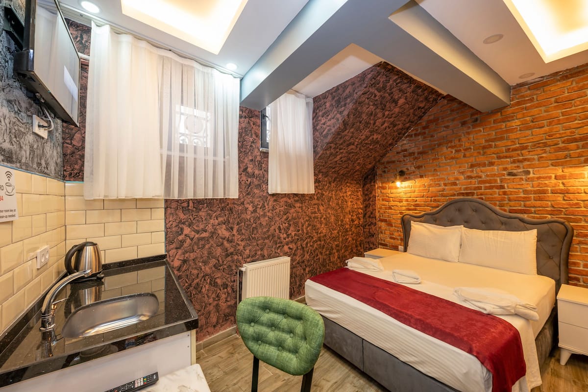 A hotel room with a brick wall, a bed, a green chair, a sink, and a window with curtains.
