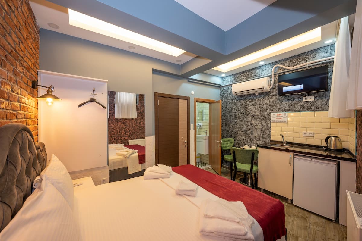 A compact hotel room with a bed, a small kitchenette, a bathroom, and a TV mounted on the wall.
