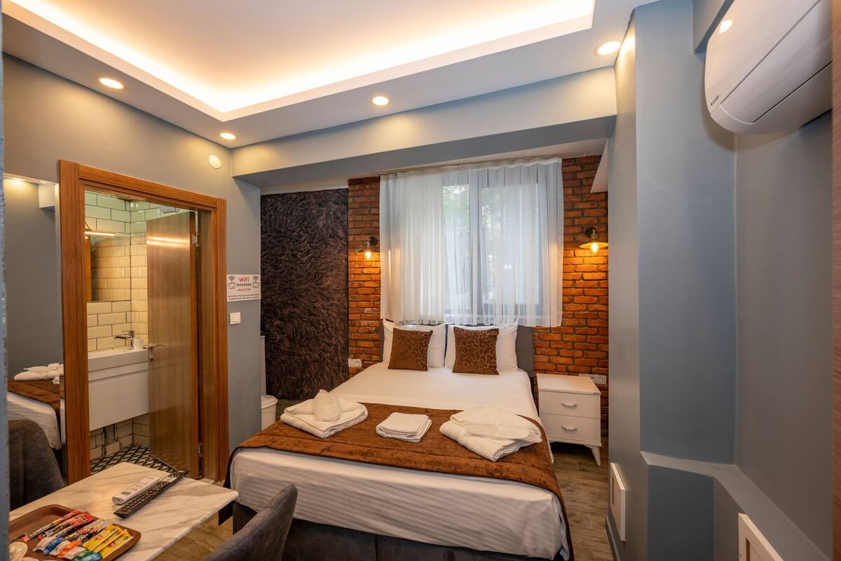 A hotel room with a bed, a desk, a chair, a mirror, a window with curtains, a brick wall, and a ceiling with recessed lighting.