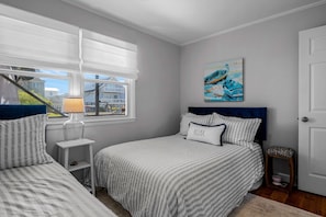 A bedroom with a bed, a nightstand, a lamp, a window with blinds, and a painting on the wall. - Charming 2BR condo in Sunset Beach, perfect for 6 guests. Enjoy the patio! (Sunset Beach)
