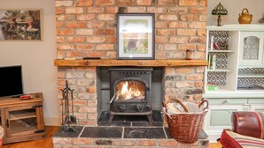 A brick fireplace with a framed picture above it, a television on a wooden stand to the left, and a wicker basket with firewood to the right.