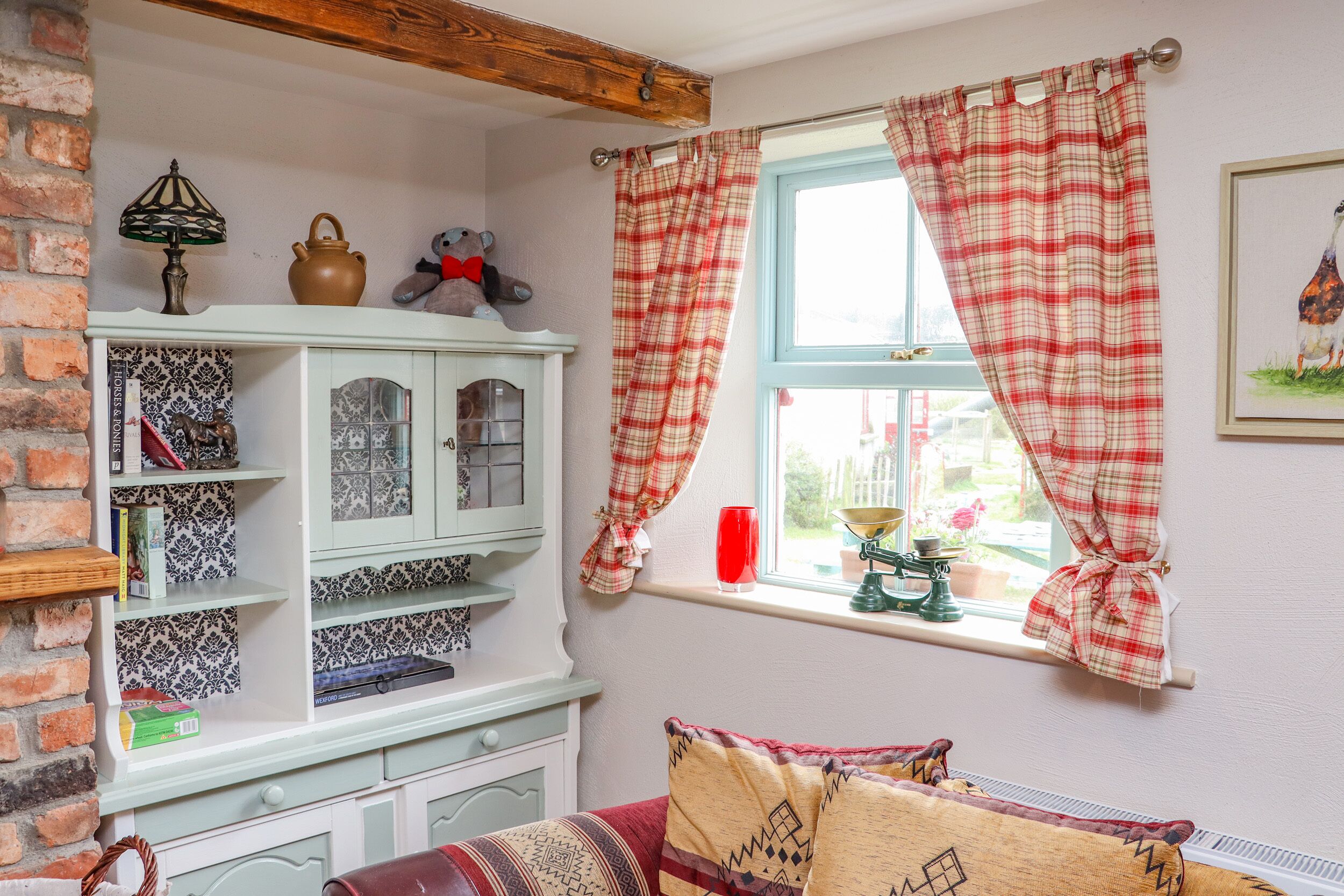 Una habitación acogedora con pared de ladrillo, un armario blanco con azulejos estampados, una ventana con cortinas a cuadros y un cojín rojo sobre un banco de madera.