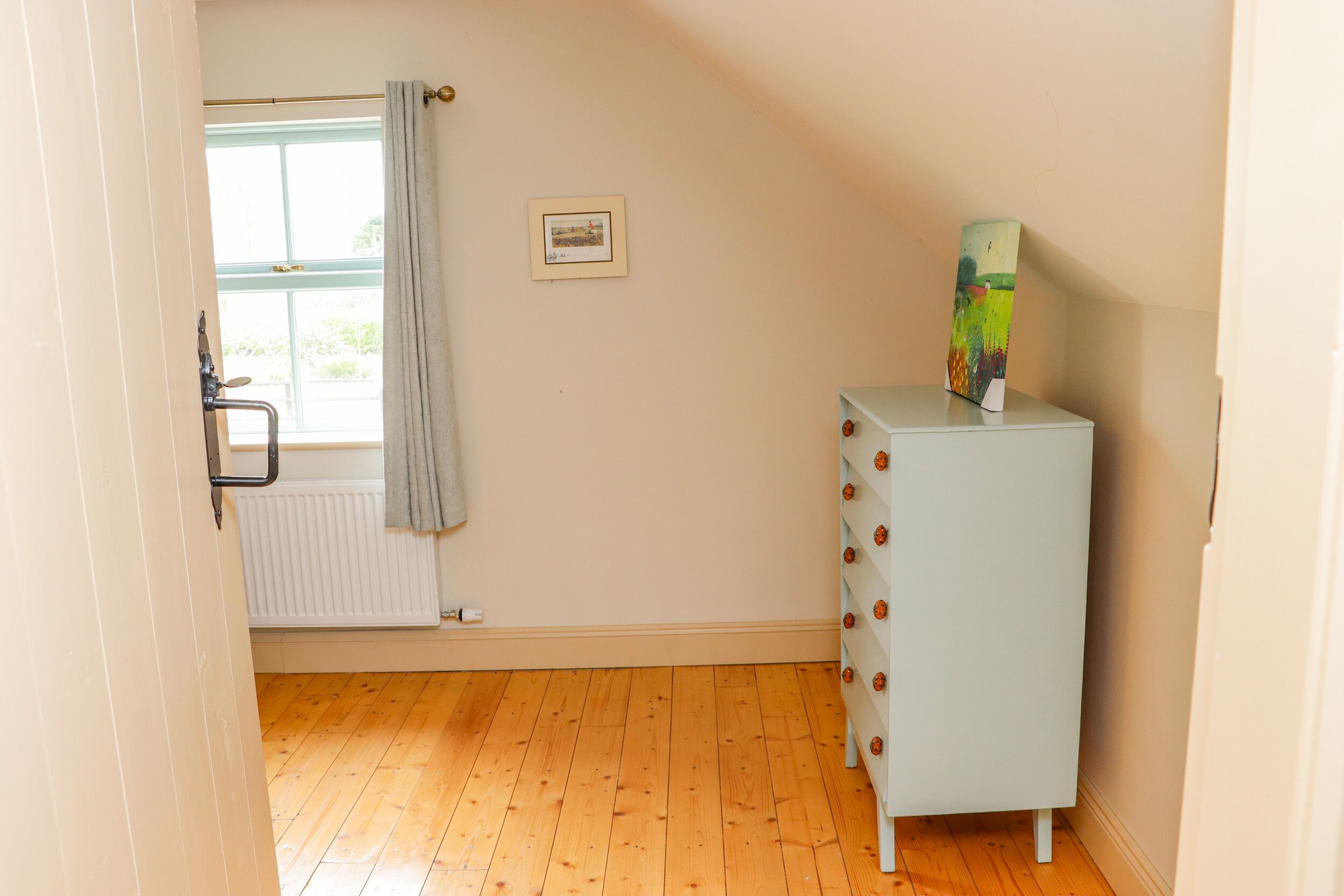 A room with a wooden floor, a radiator, a window with a curtain, and a white cabinet with a colorful object on top.