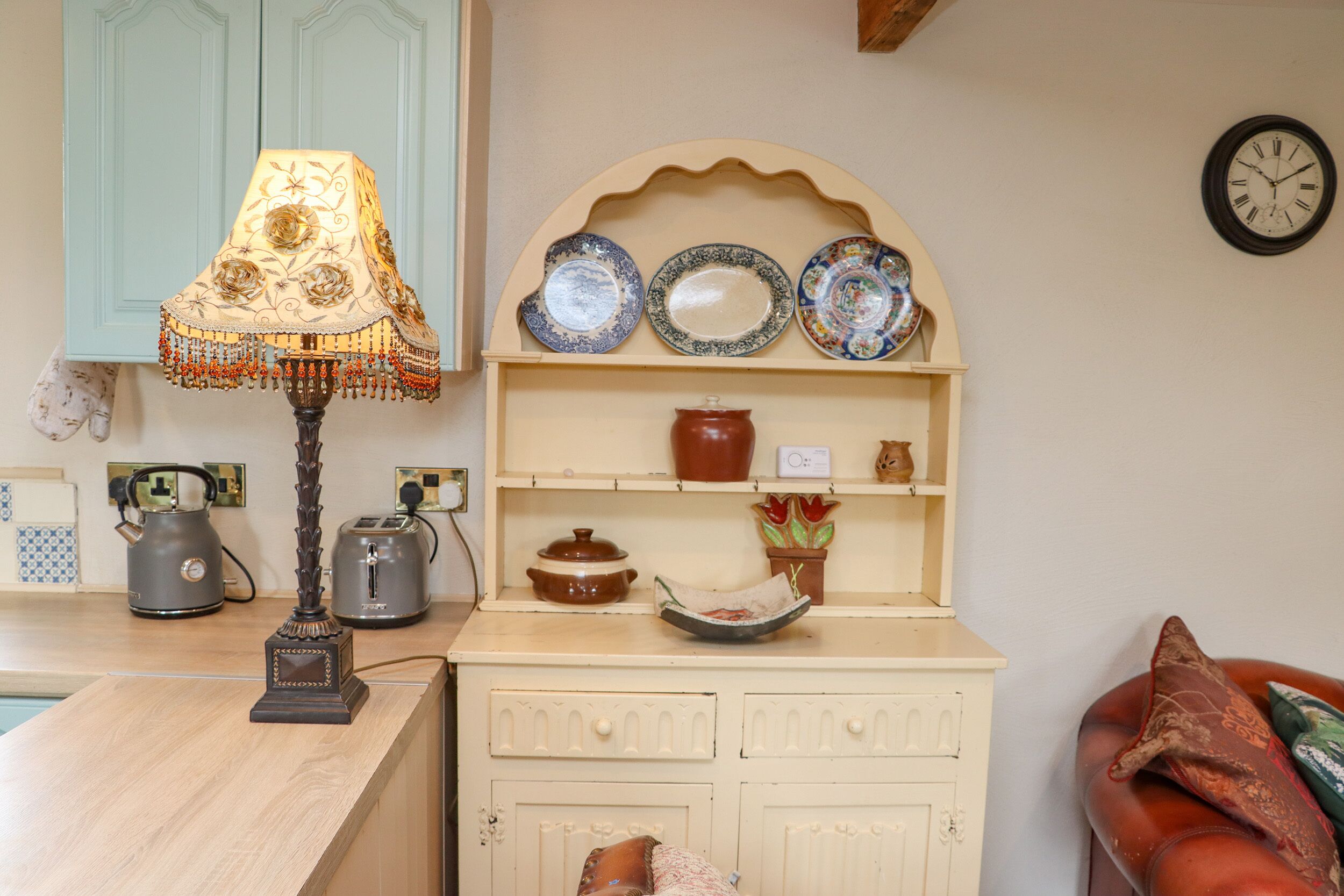 A kitchen with a wooden countertop, a decorative lamp, a shelf with plates, a clock, and a kettle.