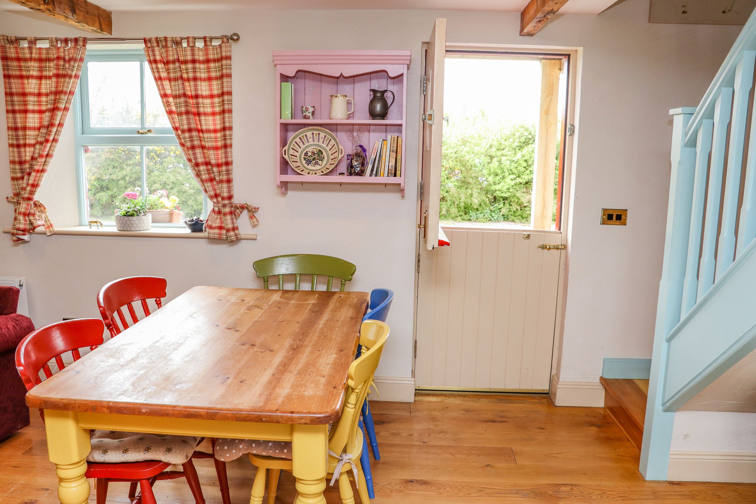 Ein Essbereich mit einem Holztisch, roten und gelben Stühlen, einem rosa Regal mit Büchern und Dekoration sowie einem Fenster mit Karottenvorhängen.