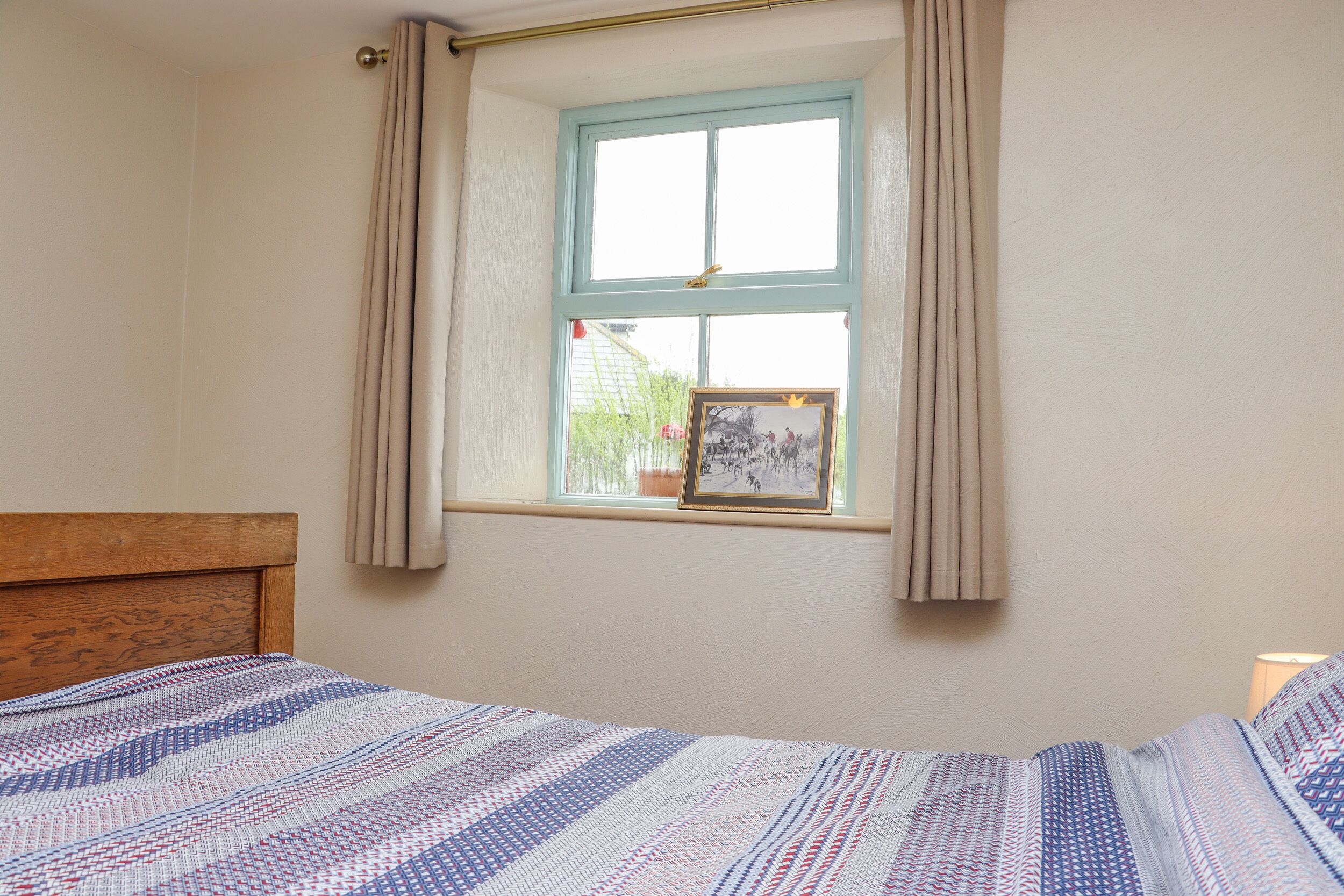 Een slaapkamer met een houten hoofdbord, een bed met een gebedrukte spreidekking, een raam met beige gordijnen en een ingelijst beeld op de vensterbank.