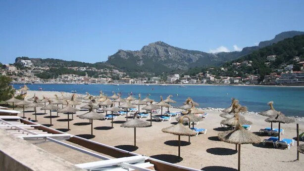 A beach with thatched umbrellas and lounge chairs, a clear blue sea, and a mountainous backdrop.