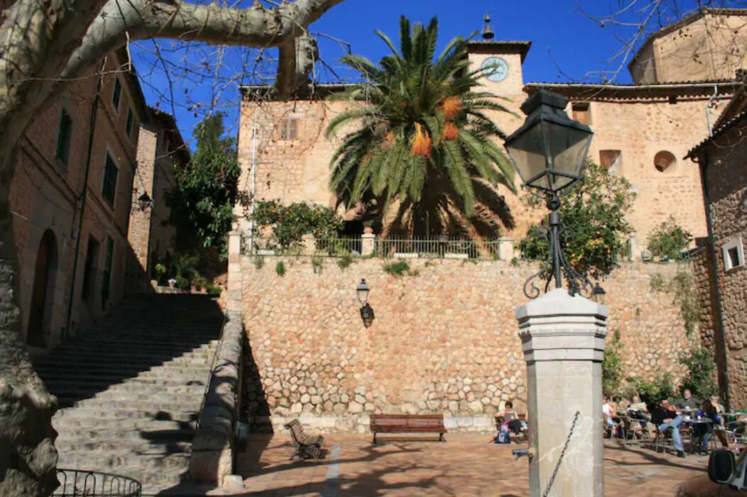 Una escalera de piedra que sube hacia un edificio con una torre de reloj, rodeado por una palmera y un muro de piedra.