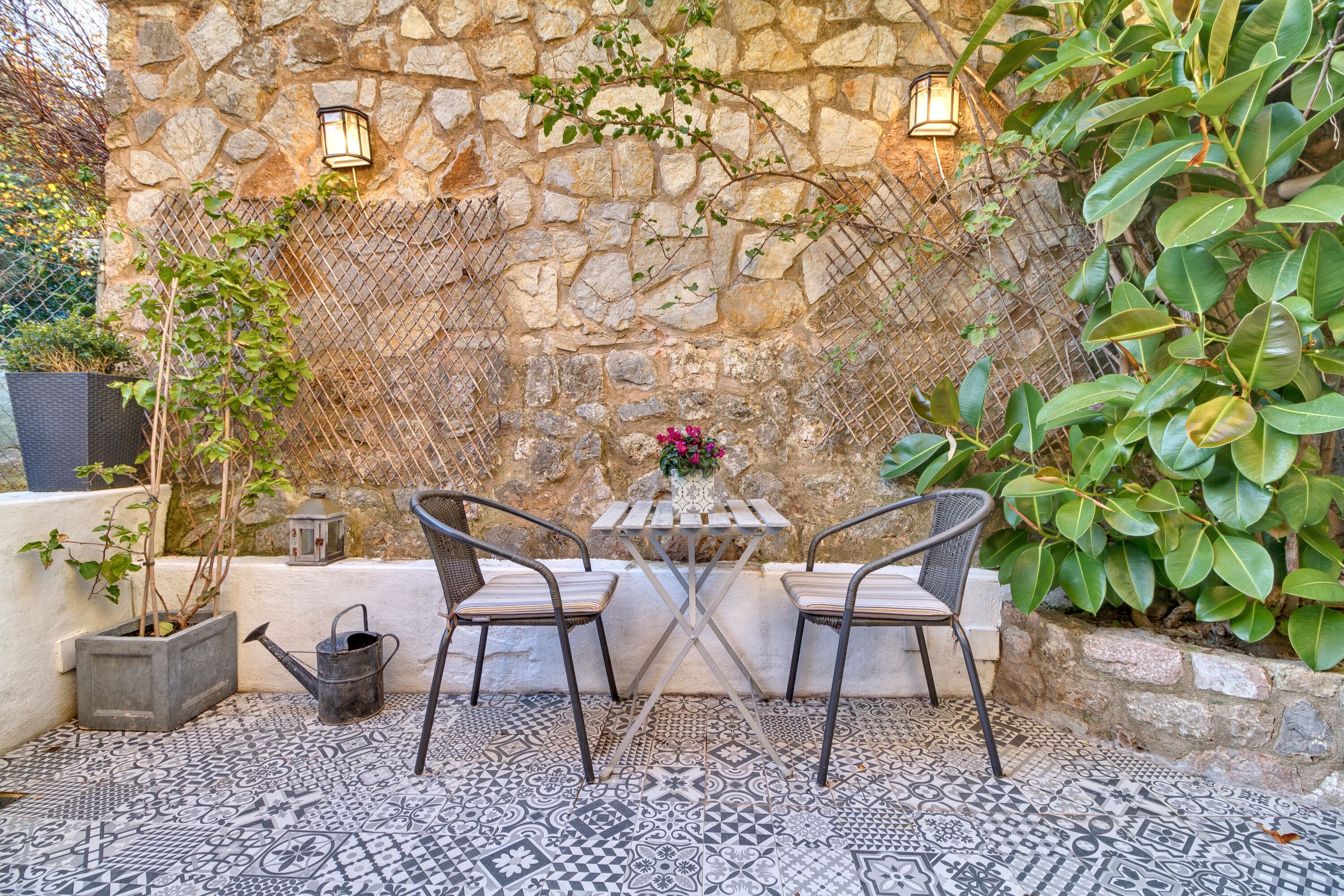 An outdoor patio with a stone wall, two metal chairs, a small table with a vase of flowers, and a tiled floor.