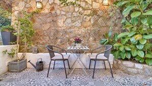 An outdoor patio with a stone wall, two metal chairs, a small table with a vase of flowers, and a tiled floor.