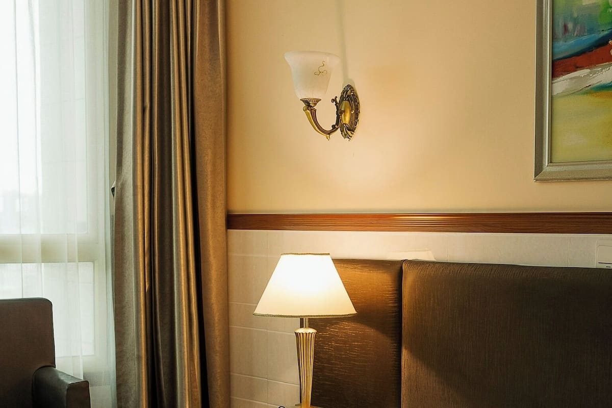 Quarto de hotel com cama, mesa de cabeceira com abajur, cadeira e um quadro na parede.