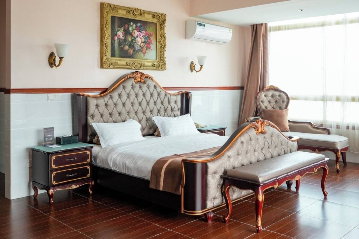 Quarto com cama grande, mesa de cabeceira, um banquinho, ar-condicionado fixado na parede e janela com cortinas.