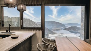 A modern kitchen with a large window, a wooden dining table, wicker chairs, and a view of mountains and a lake. - Escape View Villa Lefkada (Fterno)