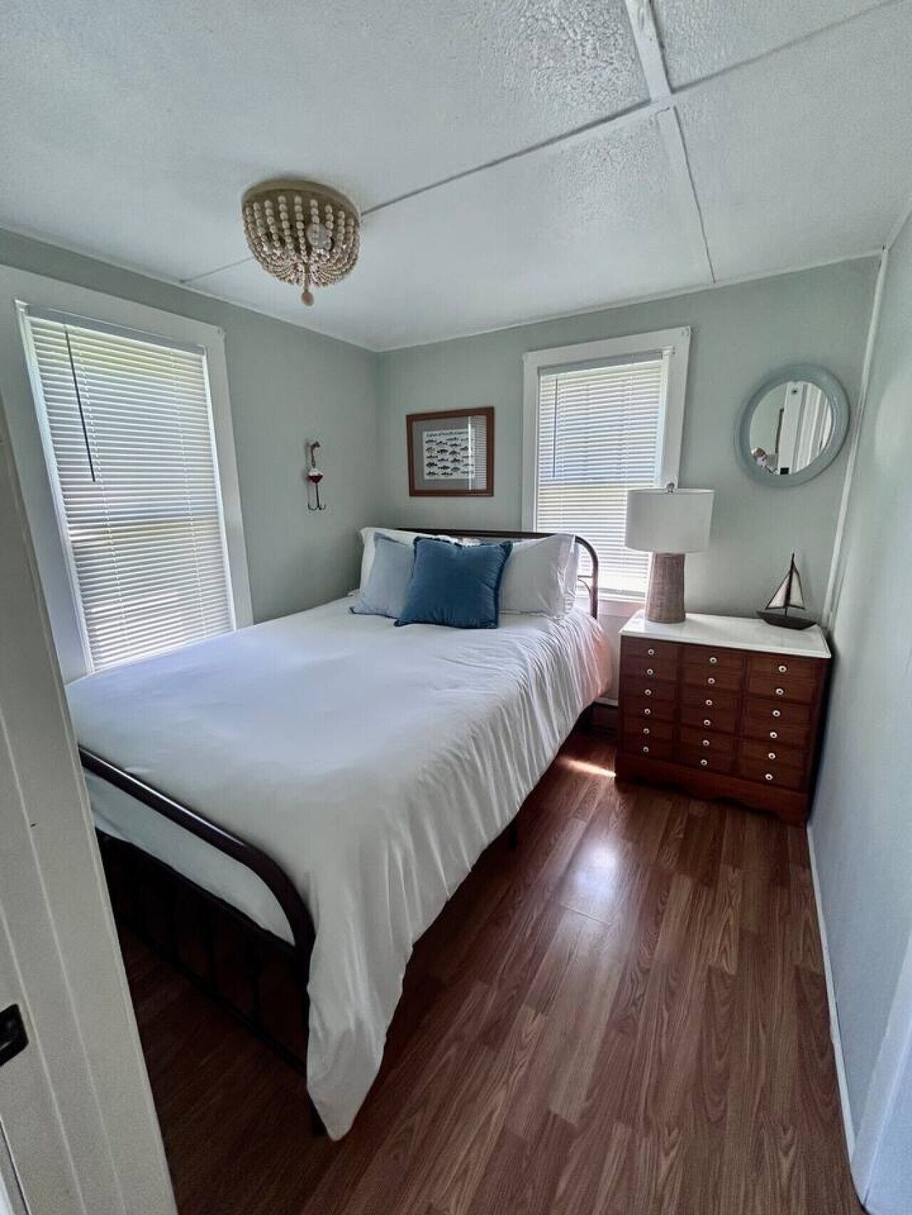 A bedroom with a bed, a dresser, a nightstand, a chandelier, and two windows with blinds.